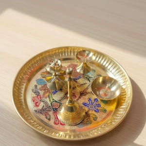 May include: A golden brass puja thali set with a floral design. The set includes a round tray, a bell, a small bowl, and two other decorative items. The tray has a detailed edge and colorful floral patterns in red, blue, and green.