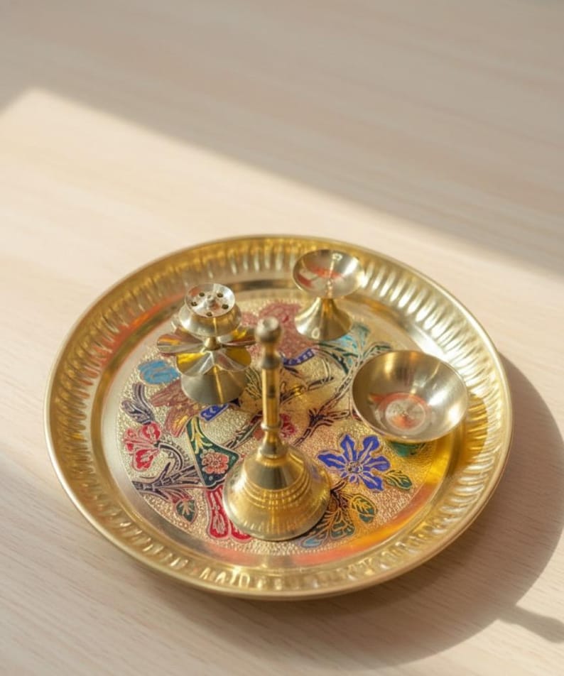 May include: A golden brass puja thali set with a floral design. The set includes a round tray, a bell, a small bowl, and two other decorative items. The tray has a detailed edge and colorful floral patterns in red, blue, and green.
