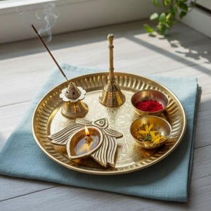 May include: A brass puja thali with a lit oil lamp, incense, and small bowls containing red powder and yellow flowers. The thali sits on a blue cloth, creating a serene and spiritual ambiance. A small bell and incense burner are also present.