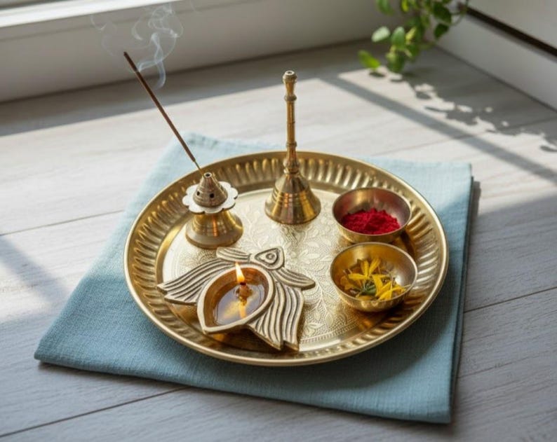 May include: A brass puja thali with a lit oil lamp, incense, and small bowls containing red powder and yellow flowers. The thali sits on a blue cloth, creating a serene and spiritual ambiance. A small bell and incense burner are also present.
