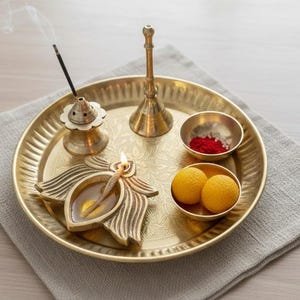 May include: A golden brass puja thali with various items for a religious ceremony. The thali holds a lit oil lamp, incense, small bowls with red powder and yellow sweets, and a bell. The items are arranged on a gray cloth.