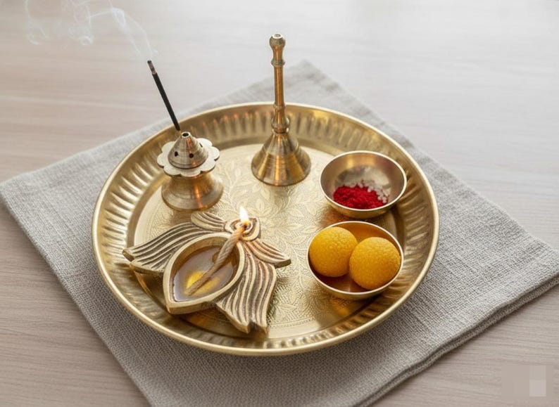 May include: A golden brass puja thali with various items for a religious ceremony. The thali holds a lit oil lamp, incense, small bowls with red powder and yellow sweets, and a bell. The items are arranged on a gray cloth.