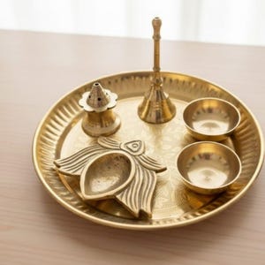 May include: A golden brass puja thali set with a decorative floral design. The set includes a round tray, two small bowls, a bell, a lamp, and an incense holder. The items are arranged on a light-colored wooden surface.