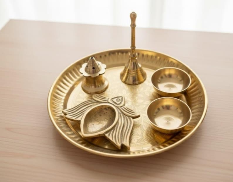 May include: A golden brass puja thali set with a decorative floral design. The set includes a round tray, two small bowls, a bell, a lamp, and an incense holder. The items are arranged on a light-colored wooden surface.