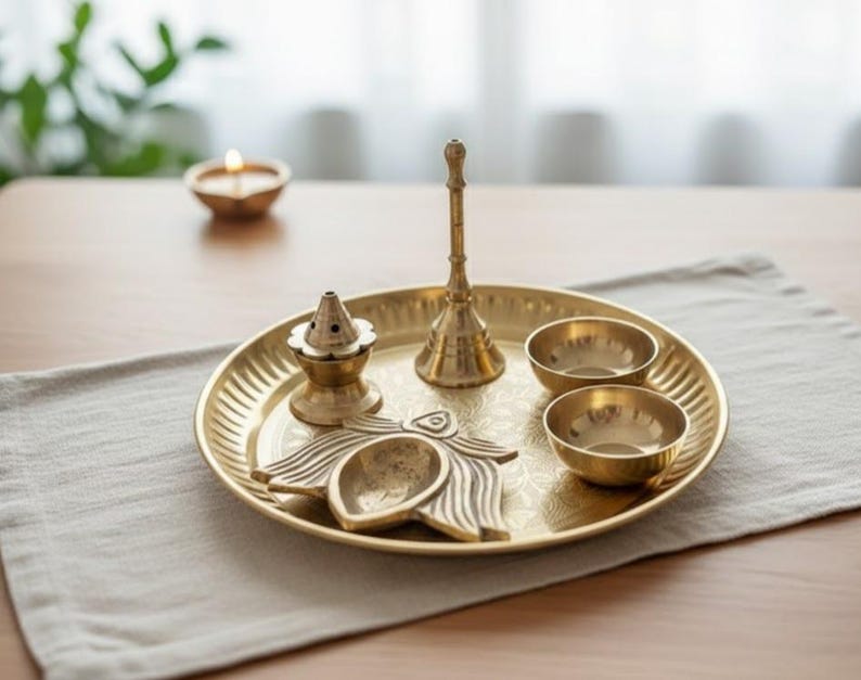 May include: A golden brass puja thali set with a bell, two small bowls, a lamp, and an incense holder. The thali is on a light gray placemat on a wooden table. A lit candle is in the background.