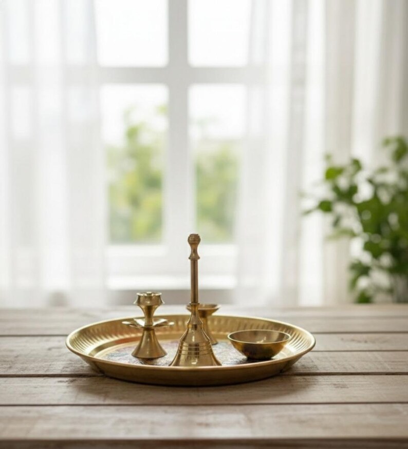 May include: A golden brass tray with decorative items, including a bell, a small bowl, and a candle holder. The items are arranged on a wooden table in front of a window with sheer white curtains. The tray is approximately 10 inches in diameter.