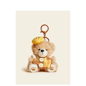 May include: A brown teddy bear wearing a yellow and brown hat holds a wooden cutting board with a painted design of eggs and bread. The bear is attached to a silver key chain.