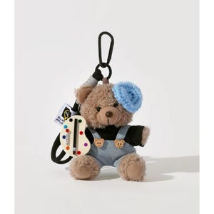 Puede incluir: Un llavero de oso de peluche marrón con boina azul y peto vaquero. El oso sostiene una paleta con puntos de colores y un mosquetón negro. El texto "key chain" está en la parte superior izquierda.
