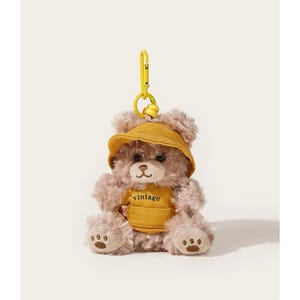 Puede incluir: Llavero de oso de peluche con sombrero amarillo y chaleco amarillo con la palabra "vintage". El llavero tiene un cierre amarillo. El texto "key chain" está en la parte superior izquierda.