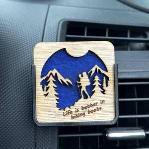 May include: A square wooden car air freshener clip featuring a laser-cut design of a hiker with a backpack among mountains and pine trees, set against a deep blue background. The text "Life is better in hiking boots" is engraved at the bottom.