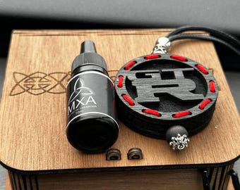 May include: A wooden box with a black bottle labeled "MXA Art of Car Aroma 10ml" and a black pendant with red stitching. The pendant has a black cord and a silver-colored bead. Two small wooden pieces are also visible.