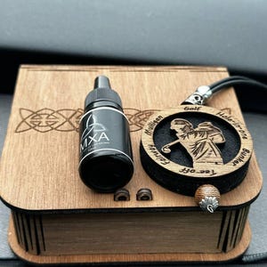 May include: A wooden box with a golf-themed pendant and a black bottle of car aroma. The pendant has a golfer silhouette and the words "Golf Hole-in-One". The bottle is labeled "MXA" and "NEOF CAR AROMA".