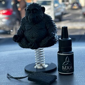 Könnte beinhalten: Eine schwarze Affenfigur auf einer Feder, eine kleine schwarze Flasche mit weißem Text und ein schwarzer rechteckiger Lufterfrischer. Die Flasche trägt den Text "MXA" und "THE ART OF CALM". Der Hintergrund ist unscharf.