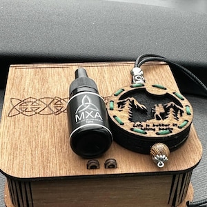 May include: A wooden box with a Celtic knot design, a black bottle labeled "MXA", and a pendant with the text "Life is better in hiking boots". The pendant features a mountain scene and green accents.