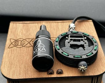 May include: A wooden box with a decorative Celtic knot design holds a black bottle labeled "MXA Car Aroma 10ml" and a black and green Jeep-shaped car air freshener. The air freshener has a black cord and a decorative bead.