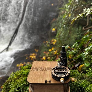 Könnte beinhalten: Eine Holzkiste mit keltischen Knoten, ein schwarzes und silbernes Nissan-Emblem und eine schwarze Flasche mit dem Text "MXA". Die Artikel werden auf grünem Moos mit einem Wasserfall im Hintergrund ausgestellt.