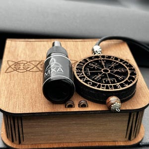 May include: A wooden box with a carved design contains a black bottle labelled "MXA Car Aroma" and a circular pendant with a rune symbol. Two small wooden pieces and a decorative bead are also present.