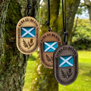 May include: Wooden pendant necklaces with the Scottish flag design. Each pendant features the flag's white saltire on a blue background, surrounded by a decorative border and the text "ALBA GU BRÁTH".