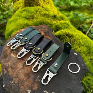 Puede incluir: Llaveros de cuero en varios colores, incluyendo verde y negro, con herrajes plateados y dorados. Los llaveros se exhiben sobre una piedra oscura, con un fondo de musgo verde.