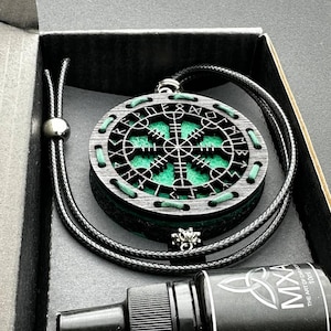 May include: A circular black wooden pendant featuring a white Vegvisir symbol and runic inscriptions, accented with green stitching and a black cord necklace. Included is a small black 10ml bottle labelled MXA The Art of Carara.