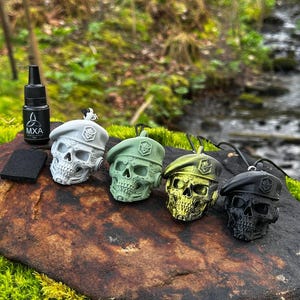 Puede incluir: Cuatro colgantes con forma de calavera en gris, verde, dorado y negro, cada uno con una boina. A la izquierda hay una botella negra con texto blanco y un rectángulo negro. Los colgantes están sobre una roca musgosa con un fondo natural borroso.