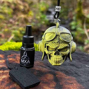 Może przedstawiać: Złoty wisiorek w kształcie czaszki z goglami lotniczymi i słuchawkami wisi na czarnym sznurku. Na zdjęciu znajduje się również czarna butelka z napisem "MXA" oraz czarny prostokątny odświeżacz powietrza. Tło to rozmyta scena na zewnątrz.