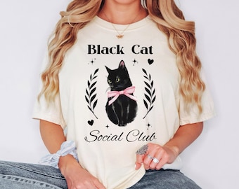 May include: Cream-colored t-shirt featuring a black cat illustration with a pink bow, surrounded by laurel leaves. The text "Black Cat Social Club" is printed above and below the cat design.