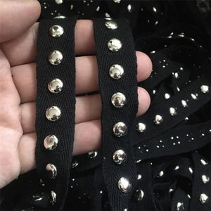 Può includere: Primo piano di nastro nero impreziosito da borchie metalliche argentate lucide. Il nastro è tenuto in mano, con altro nastro visibile sullo sfondo. Le borchie sono uniformemente distanziate lungo il nastro.