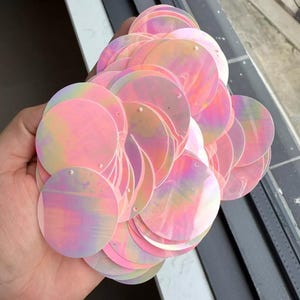 May include: A collection of iridescent, round sequins in shades of pink, peach, and lavender. Each sequin has a small hole for stringing. The sequins are held in a hand, and the light reflects off the surfaces.