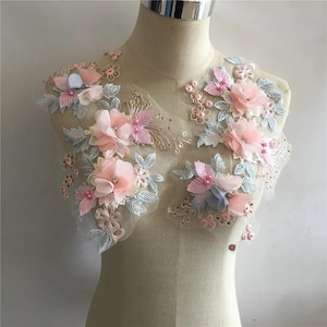 May include: Floral applique with pink, blue, and gold accents. The applique features delicate fabric flowers, leaves, and beaded embellishments. The design is on a sheer, light-colored mesh backing, perfect for embellishing garments or crafts.