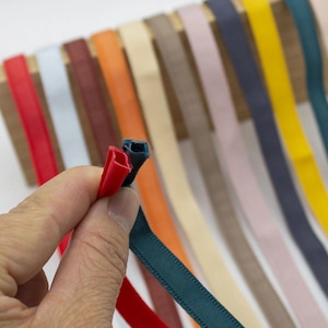 Op de afbeelding: Een hand houdt drie kleurrijke linten vast: rood, donkerblauwgroen en blauwgroen. De achtergrond toont een verscheidenheid aan linten in rood, lichtblauw, bruin, oranje, beige, roze, marineblauw, geel en blauwgroen, gepresenteerd.