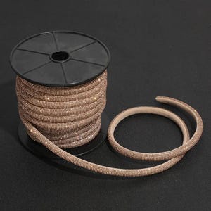 Puede incluir: Un carrete de adorno de diamantes de imitación en oro rosa con una superficie densa y brillante de pequeños cristales reflectantes. El adorno está enrollado alrededor de un carrete de plástico negro, con una parte del material flexible extendida.