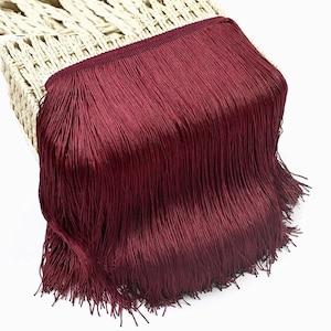 May include: A deep burgundy fringe trim featuring a dense row of long, straight, silky threads attached to a woven header band. The fringe hangs vertically in a uniform, smooth drape, showcasing a consistent dark red color throughout.