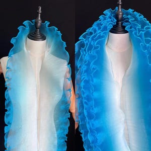 Può includere: Sciarpa blu sfumata con bordo a balze, esposta su un manichino. Il tessuto trasparente passa da un colore chiaro al centro a un blu vibrante sui bordi. Il bordo a balze aggiunge consistenza e interesse visivo.