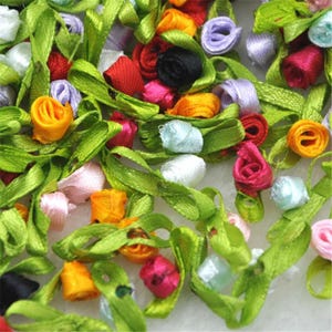 Puede incluir: Una colección de pequeñas rosas de cinta multicolores en tonos rojo, naranja, amarillo, rosa, morado, negro y blanco, cada una unida a un tallo de cinta verde brillante con hojas, creando un arreglo denso y texturizado de flores de tela.