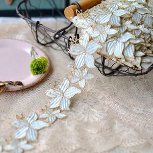 Puede incluir: Una tira continua de encaje floral blanco con motivos de flores de cinco pétalos delineados con hilo dorado metálico. El encaje está dispuesto en un patrón repetitivo, con un carrete de la cinta descansando en una cesta de alambre oscuro.