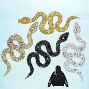 Puede incluir: Cuatro parches con forma de serpiente cubiertos de brillantes strass en dorado, multicolor, negro y plateado, cada uno con una piedra de color en el ojo. Se muestra una sudadera negra con capucha con un parche de serpiente dorada en la espalda.