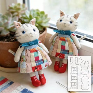 May include: Two handmade cat dolls with white bodies, gray striped heads, and blue eyes. They wear patchwork dresses, blue bow ties, and red shoes. A pattern for the dolls is included.