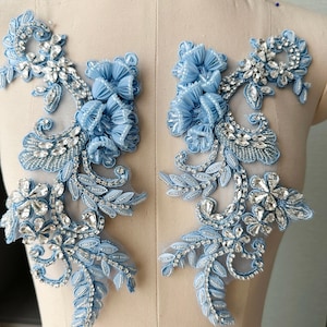 May include: A pair of symmetrical light blue floral appliques featuring intricate embroidery, pleated fabric flower petals, and clear faceted crystal rhinestones set on a sheer mesh backing, designed for decorative garment embellishment.