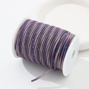 May include: A spool of purple, lavender, and silver rhinestone trim. The trim is tightly wound on a white spool. The rhinestones are small and evenly spaced, creating a sparkling effect. This trim is suitable for various craft projects.