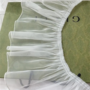 May include: A sheer white pleated tulle fabric trim featuring a gathered edge with visible white stitching. The delicate, semi-transparent material is arranged in soft, vertical folds, showcasing a lightweight and airy texture throughout.