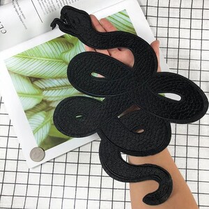 May include: A black embroidered snake patch featuring a coiled, serpentine design with a detailed head, open mouth, and visible fangs. The patch has a textured, stitched surface and a defined border, showcasing a continuous, winding body shape.