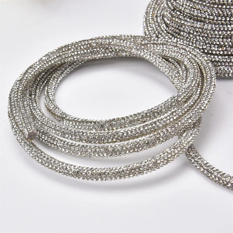May include: Coiled silver rhinestone trim. The trim is made of small, sparkling rhinestones set closely together, creating a continuous, shimmering band. The trim is flexible and appears to be on a roll, suitable for various craft and design projects.