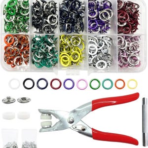 Puede incluir: Un organizador de plástico transparente con diez compartimentos llenos de broches metálicos de colores. Debajo hay diez anillos de colores, dos piezas de broche, dos almohadillas de goma blancas, una pinza de metal con mango rojo y una varilla.