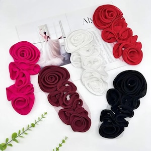 May include: An assortment of fabric flower embellishments in various colors, including red, white, black, and burgundy. The flowers are arranged on a white surface, with a magazine page visible in the background. The flowers are likely for crafting or fashion.