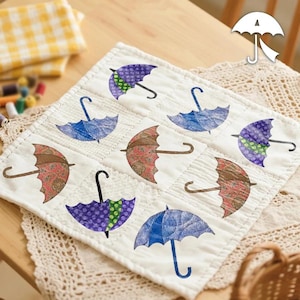 Op de afbeelding: Een wit gequilt vierkant met negen kleurrijke paraplu-ontwerpen. De paraplu's zijn in tinten blauw, paars en bruin, met zwarte gebogen handvatten. De achtergrond is lichtbeige en de algehele esthetiek is handgemaakt.