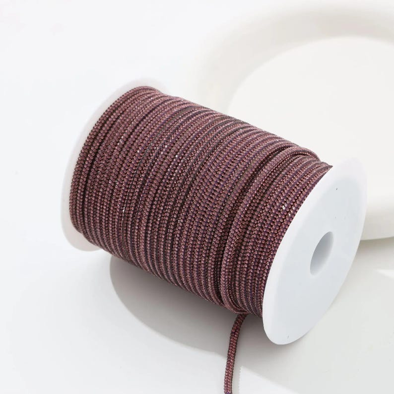 May include: A spool of mauve-colored rhinestone trim. The trim is tightly wound on a white plastic spool. The rhinestones are small and closely set, creating a continuous, sparkling surface. The trim is suitable for various craft and design projects.