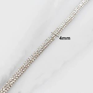 May include: Close-up of a silver rhinestone trim, 4mm wide. The trim is composed of multiple rows of small, sparkling rhinestones, creating a shimmering effect. The background is a light, marbled surface.