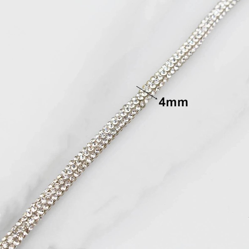 May include: Close-up of a silver rhinestone trim, 4mm wide. The trim is composed of multiple rows of small, sparkling rhinestones, creating a shimmering effect. The background is a light, marbled surface.