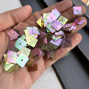 May include: A collection of small, square-shaped sequins with a reflective, iridescent finish that shifts between shades of pink, purple, green, and gold. Each individual sequin features a single, centred hole for attachment, creating a shimmering effect.
